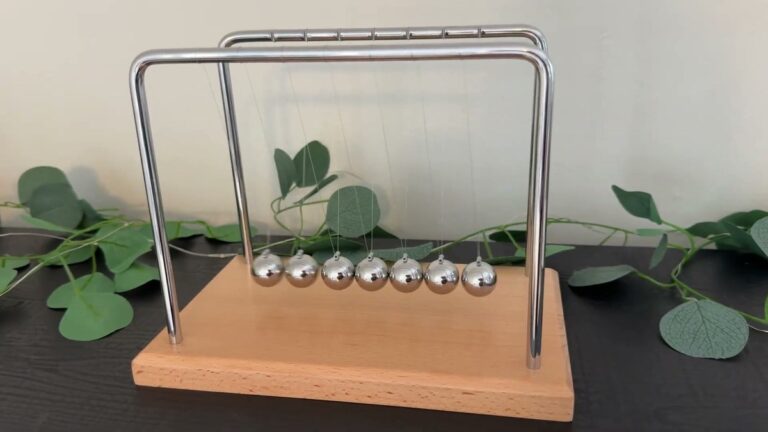 Smilesorg Newton's cradle with seven chrome balls hanging at rest from a curved steel frame on a light wood base
