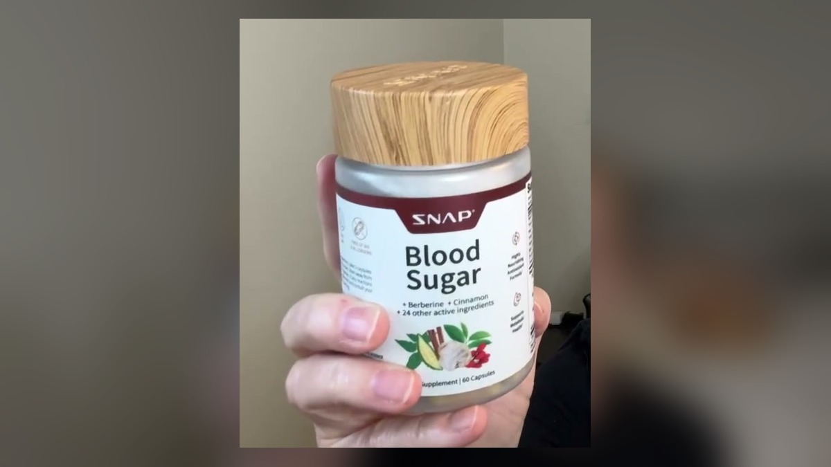Hand holding a Snap Blood Sugar supplement bottle with a wood-look lid and a white label listing berberine, cinnamon and 24 other active ingredients