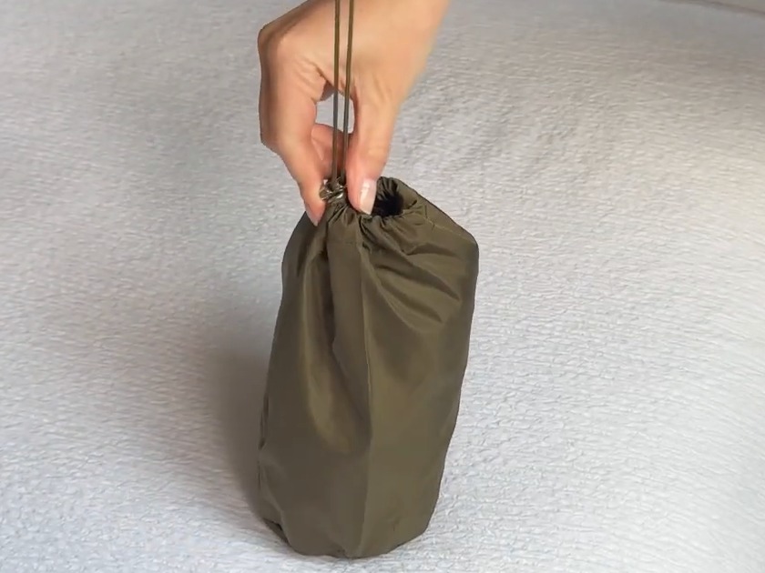 TCOT rain jacket packed into its army green drawstring pouch, held up by the cord over a white bedspread