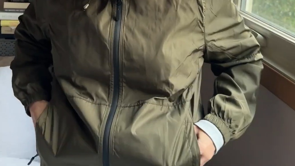 TCOT rain jacket in army green zipped up, hands tucked into the zippered side pockets