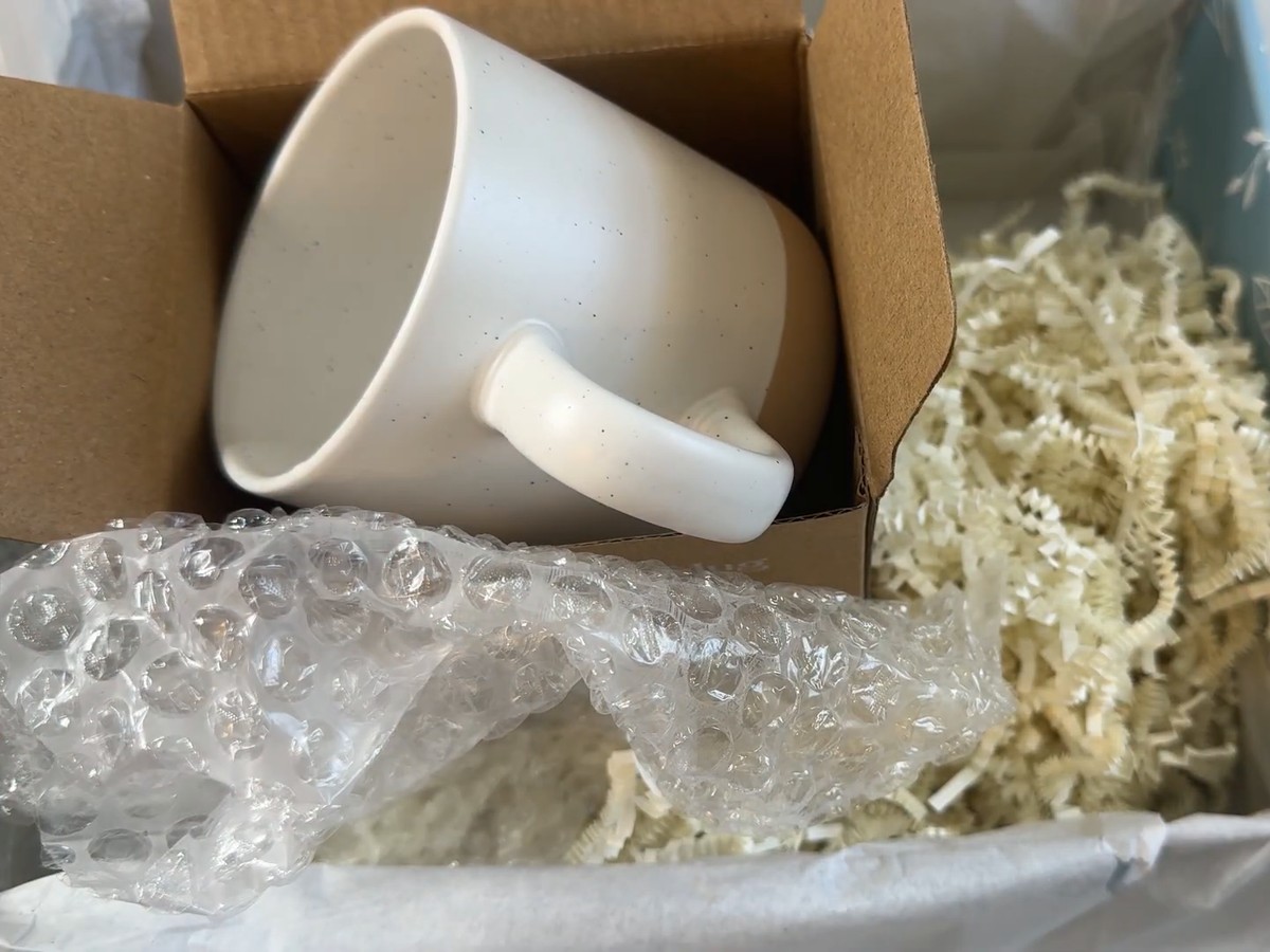 Speckled cream ceramic mug with a bare tan clay base, sitting in its open cardboard box inside the Unboxme gift basket.