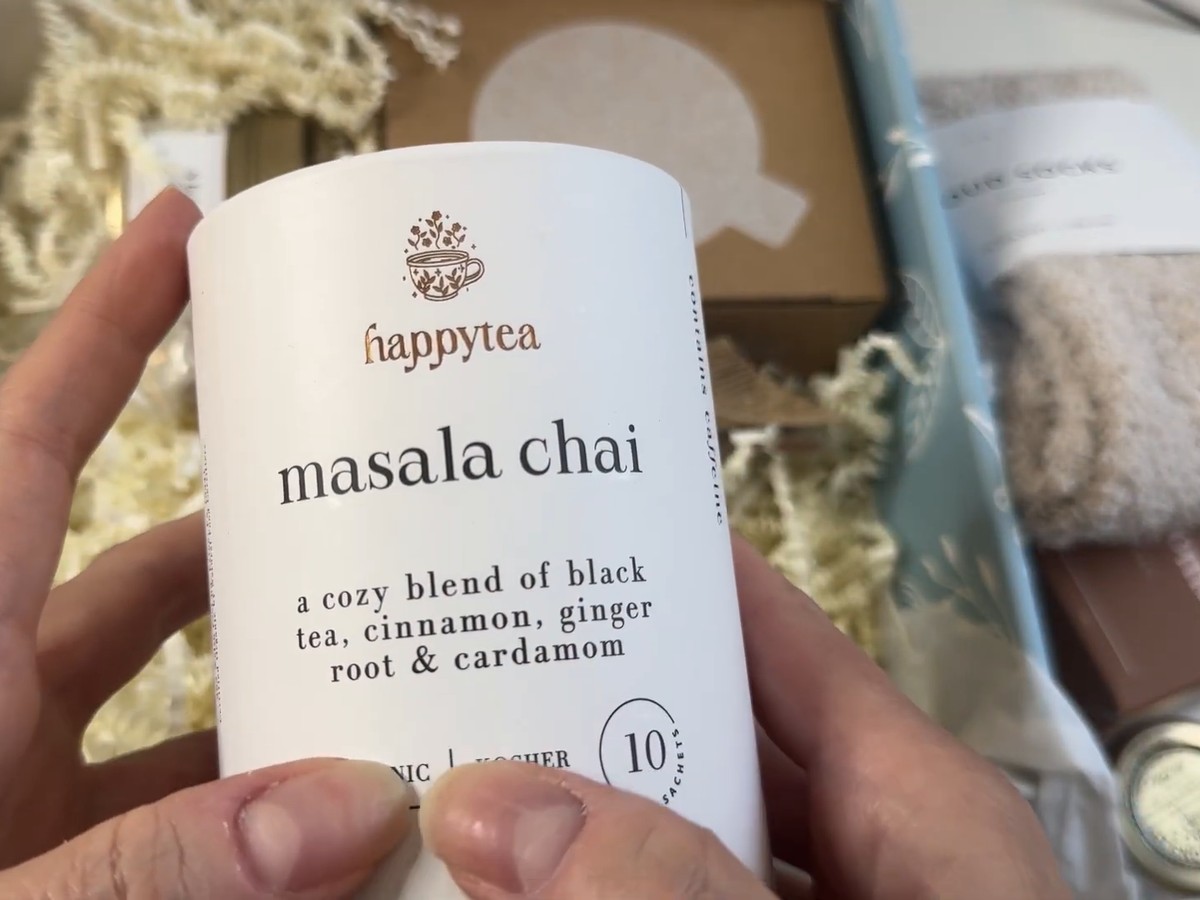 Hands holding a white happytea masala chai tin labeled as a blend of black tea, cinnamon, ginger root and cardamom with 10 sachets.