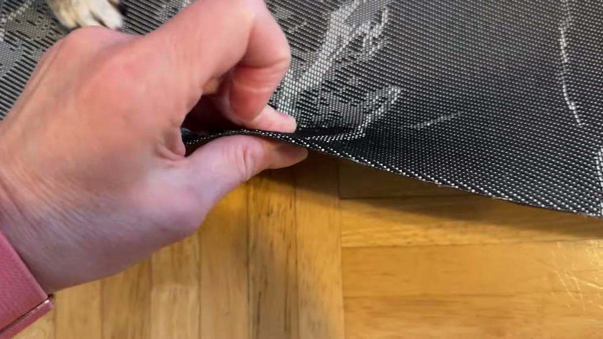 Hand lifting the corner of the Vaukki PVC chair mat to show how thin and flexible it is above the hardwood floor