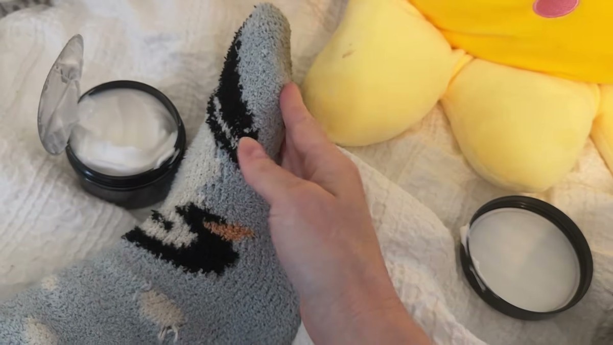 Foot in a gray and black fluffy sock on a bed next to an open tub of VREKEF urea cream