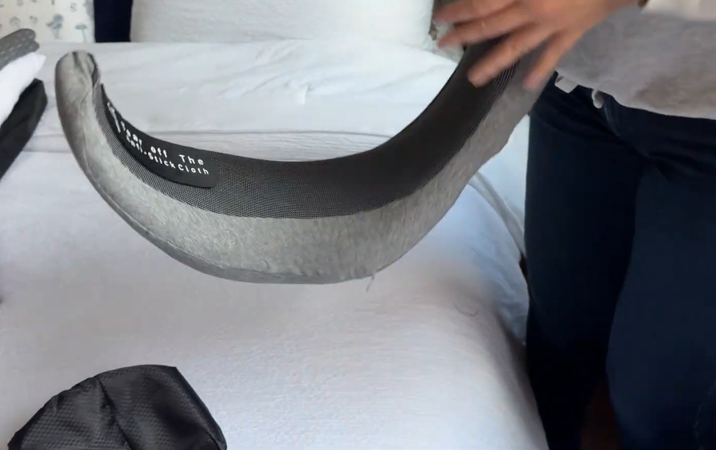 Gray WEERSHUN travel pillow held open over a bed, showing the black strip labeled to tear off the anti-stick cloth before use