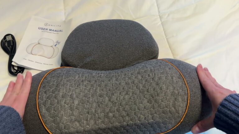 Grey Zenvita neck massage pillow with an orange-trimmed textured neck panel, lying on a bed next to its user manual and charging cord