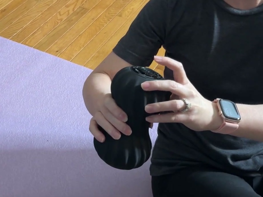Two hands holding the black Zyllion ZMA-30 peanut massage ball upright, showing its ridged silicone sides and the button panel on top